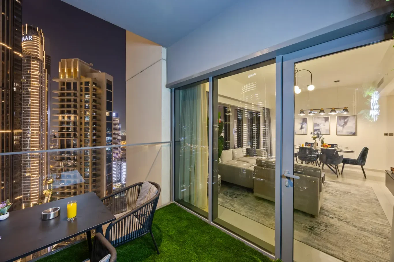 Night cityscape from a balcony with a black dining table, candles, and wicker chairs; glass sliding doors open to a modern living room.