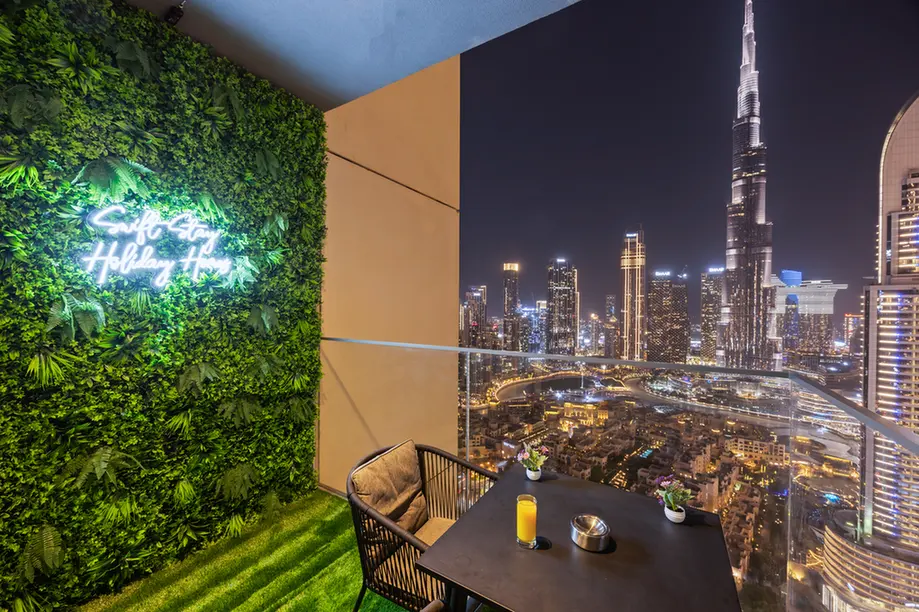 Night rooftop balcony overlooking a brightly lit city skyline; a table with a candle and small plants, two chairs, and a green plant wall with a neon sign.