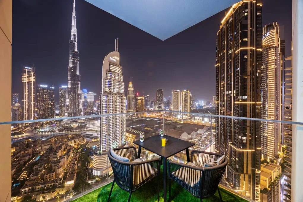 A nighttime balcony with a small table and two chairs overlooking a brightly lit city skyline and glass railing.