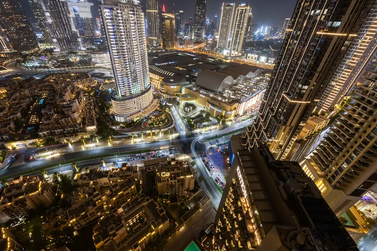 Nighttime aerial view of a dense cityscape: tall, illuminated skyscrapers, glowing streets, and a large curved-roof complex at the center.