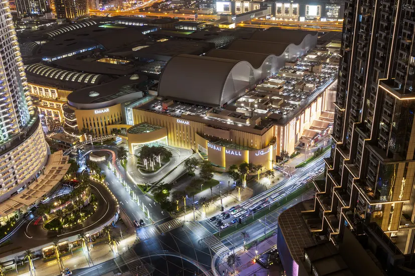 Aerial nighttime view of Dubai Mall with curved white canopies and surrounding high-rise buildings, illuminated streets.