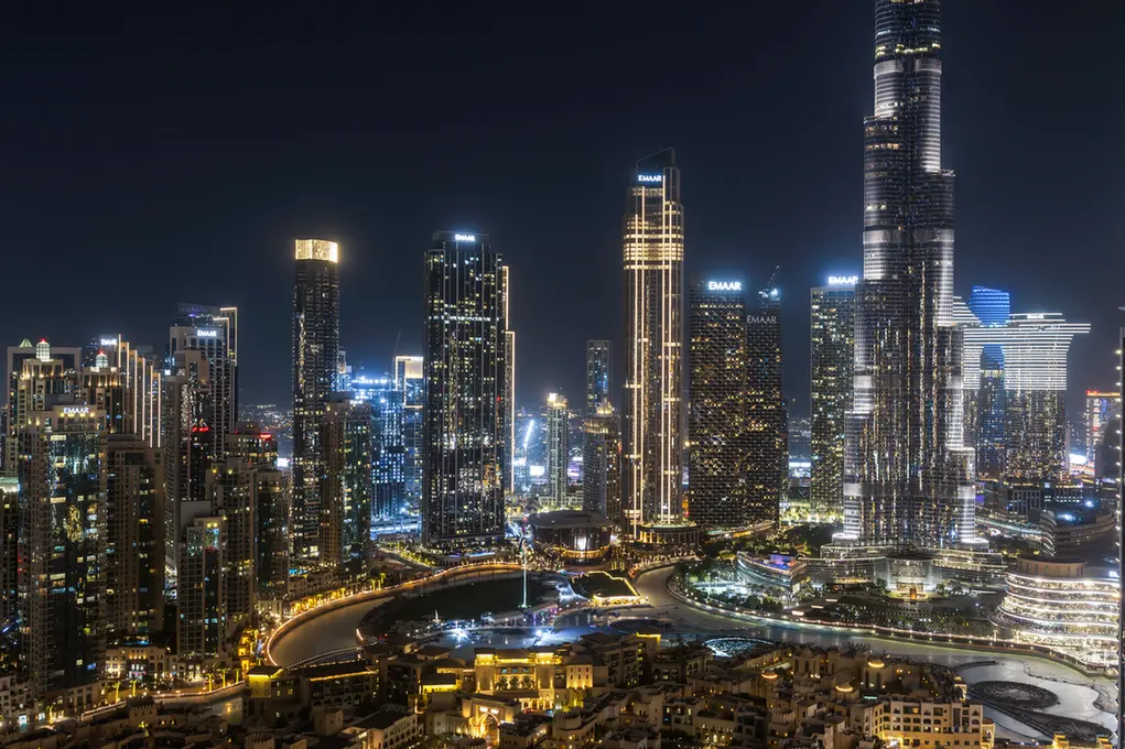 Nighttime city skyline with tall, illuminated skyscrapers and a winding road in the foreground.