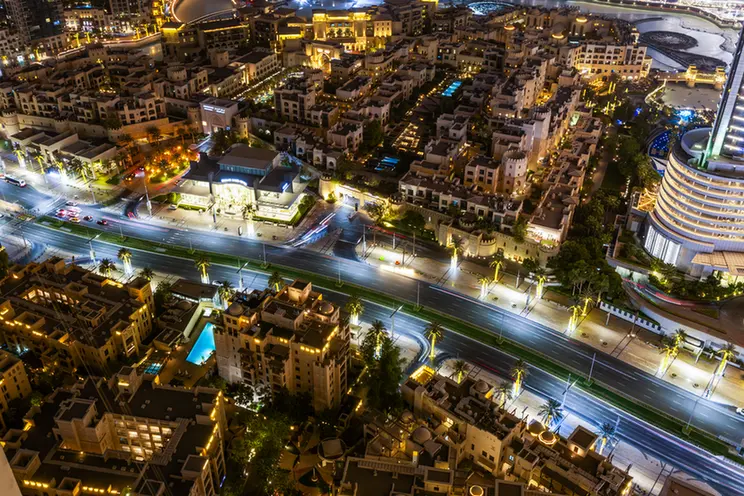 Aerial night view of a dense urban area with brightly lit streets, palm trees, and a large curved building on the right edge.