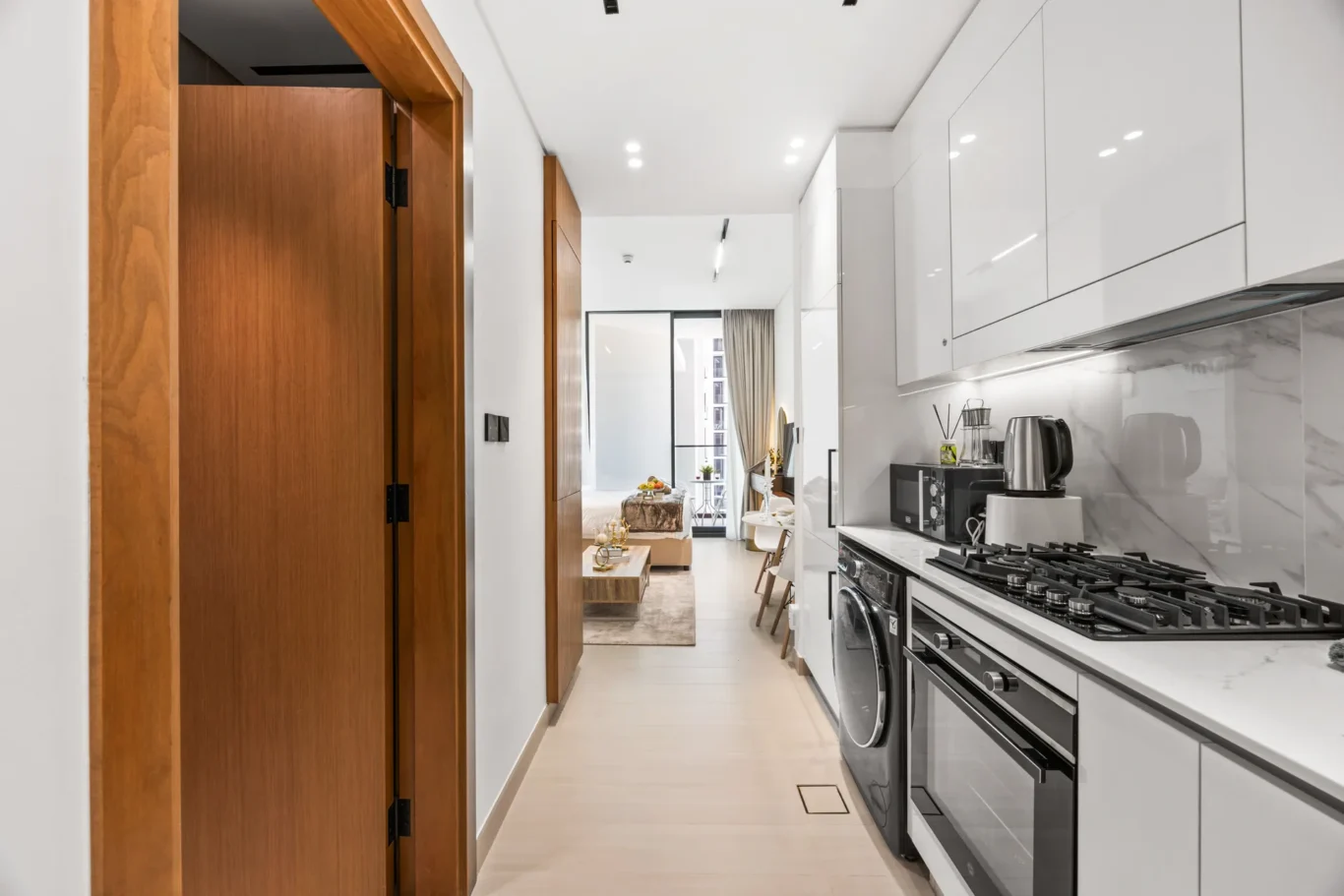 Bright contemporary kitchen with white glossy cabinets, marble backsplash, and stainless steel appliances, opening to a living area.