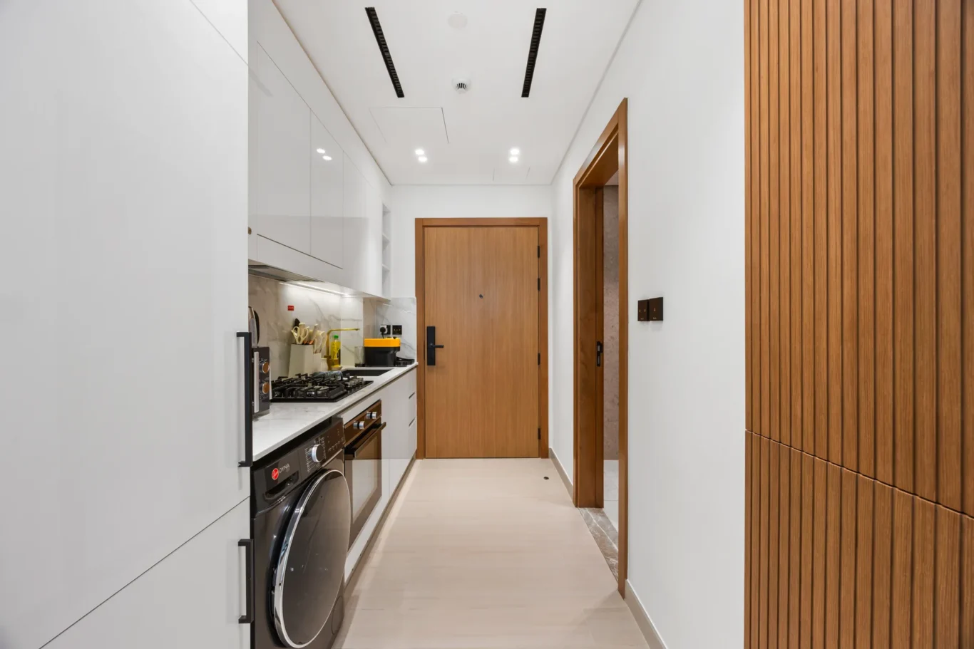 Narrow modern kitchen hallway with white glossy cabinets, built-in appliances, and a wooden door at the end, plus a vertical wood-paneled wall on the right.