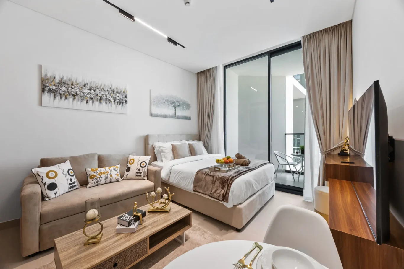 Modern hotel-suite living area with beige sofa, bed, and large glass balcony doors; neutral decor and abstract wall art.