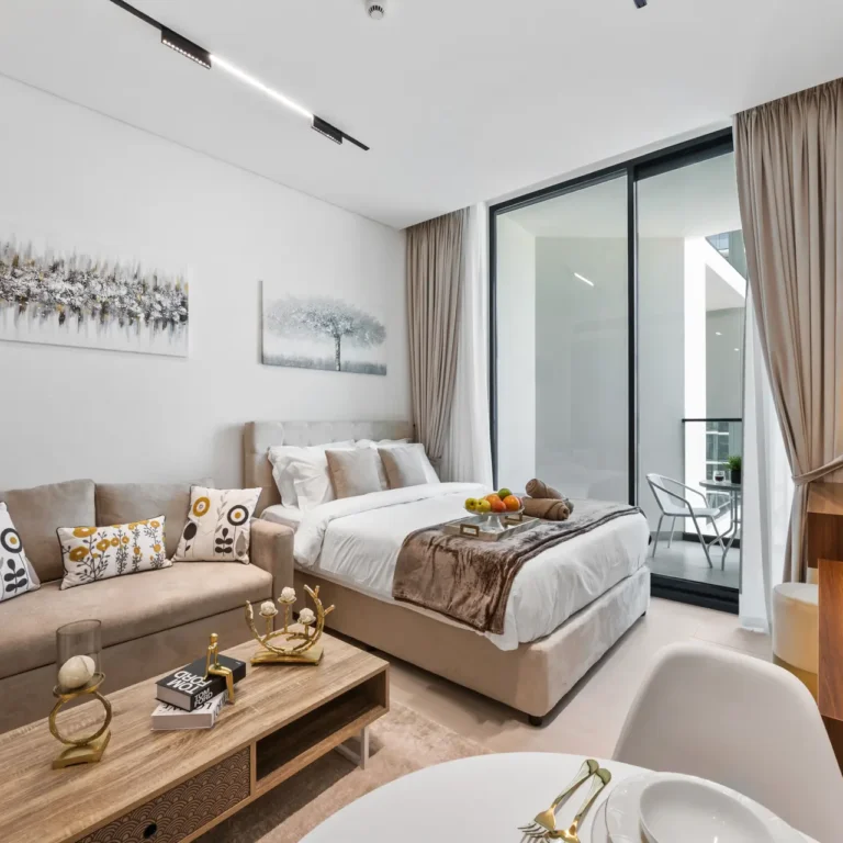 Modern hotel-suite living area with beige sofa, bed, and large glass balcony doors; neutral decor and abstract wall art.