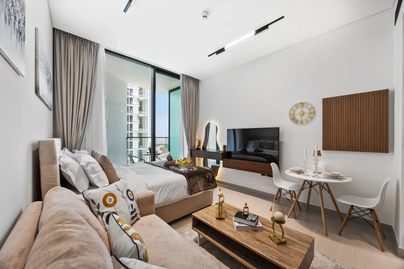 Open-plan living area with a bed, beige sofa, and wood coffee table facing a wall-mounted TV