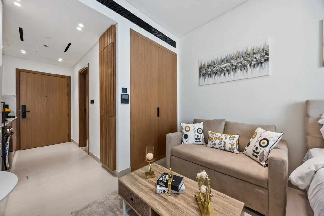 Modern living room with a beige sofa, patterned cushions, and abstract wall art on white walls with wooden doors nearby.