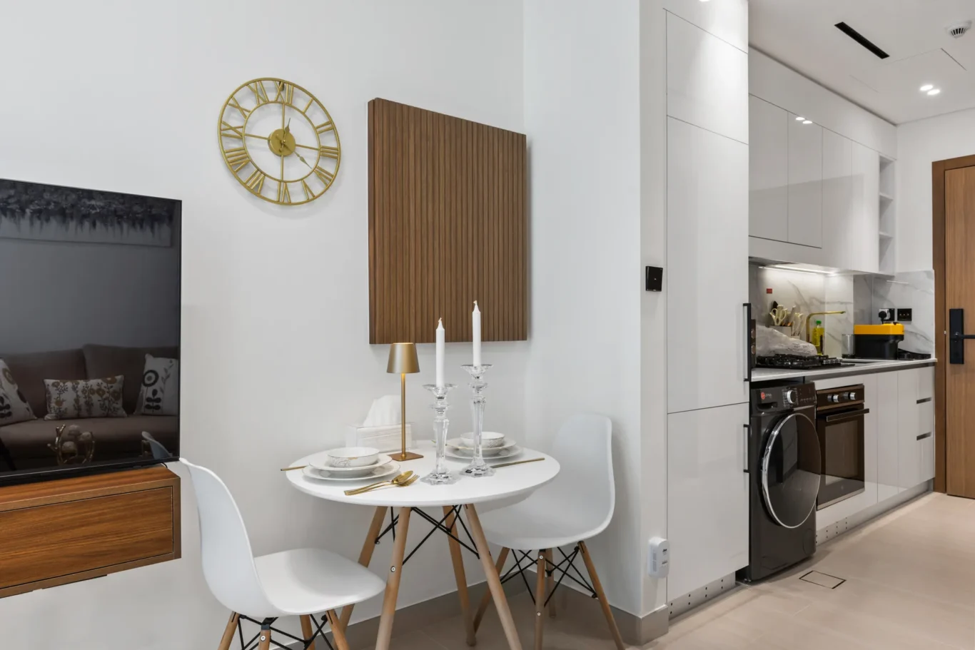 Round white dining table set for two with gold flatware and candlesticks, white chairs, in a modern kitchen and living area with a wooden wall panel and gold clock above the sofa TV area nearby.