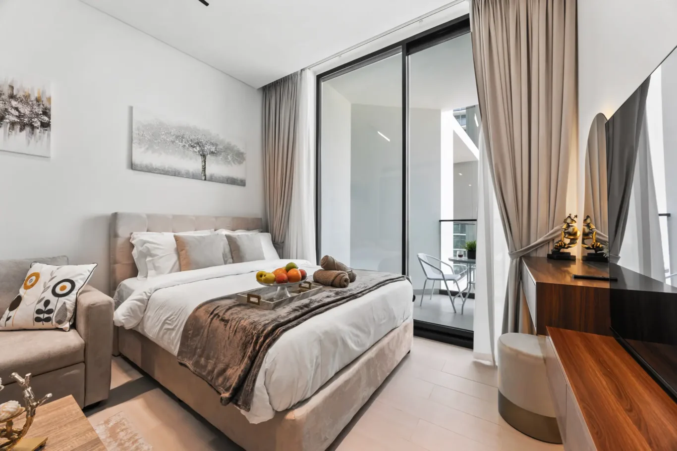 Modern hotel bedroom with a large bed, beige upholstery, and multiple pillows; sliding glass doors open to a balcony with sheer curtains. A desk with two gold statues sits to the right, and a fruit tray and towels rest on the bed. Artwork of trees hangs on the white walls.