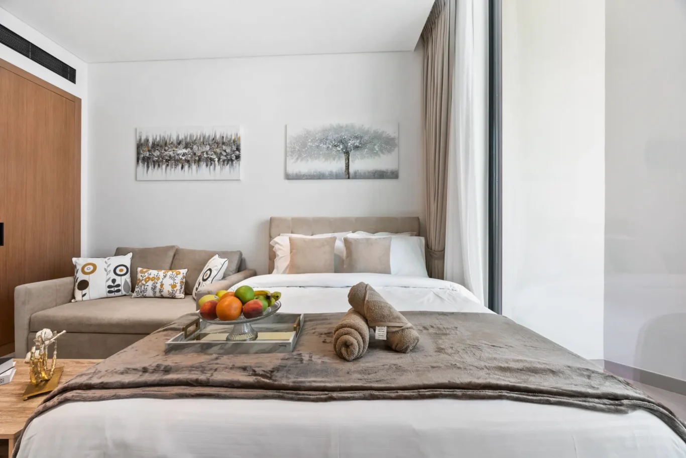 Bright hotel bedroom with a large bed, beige headboard, decorative pillows, and a fruit bowl on a tray at the foot of the bed.