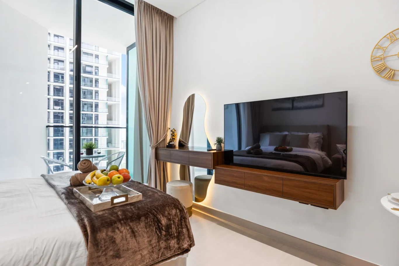 Modern hotel room interior with a bed, fruit tray, and a brown throw on a white bedspread; large window with a balcony and city buildings outside.