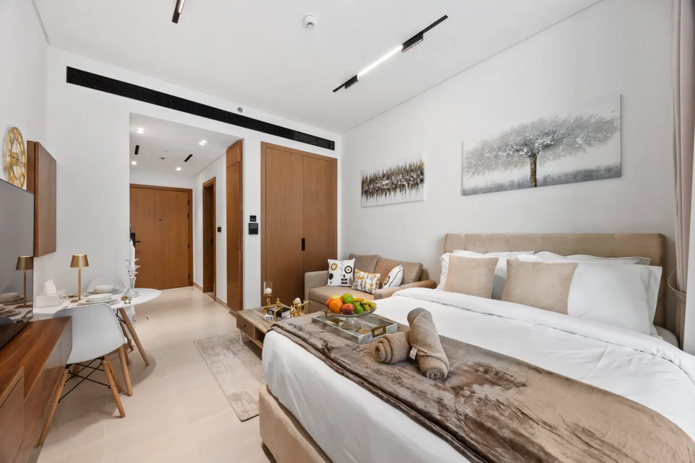 Hotel suite living area with a large bed, beige headboard, sofa, and coffee table featuring fruit and decorations; wall art above.