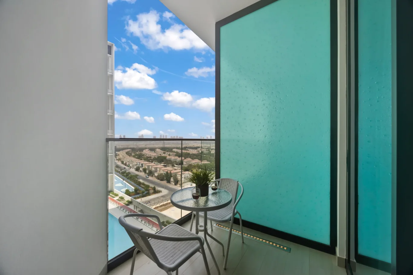 Balcony seating area with turquoise glass panels, a round glass table, two chairs, and a city view beyond the railing under a blue sky.