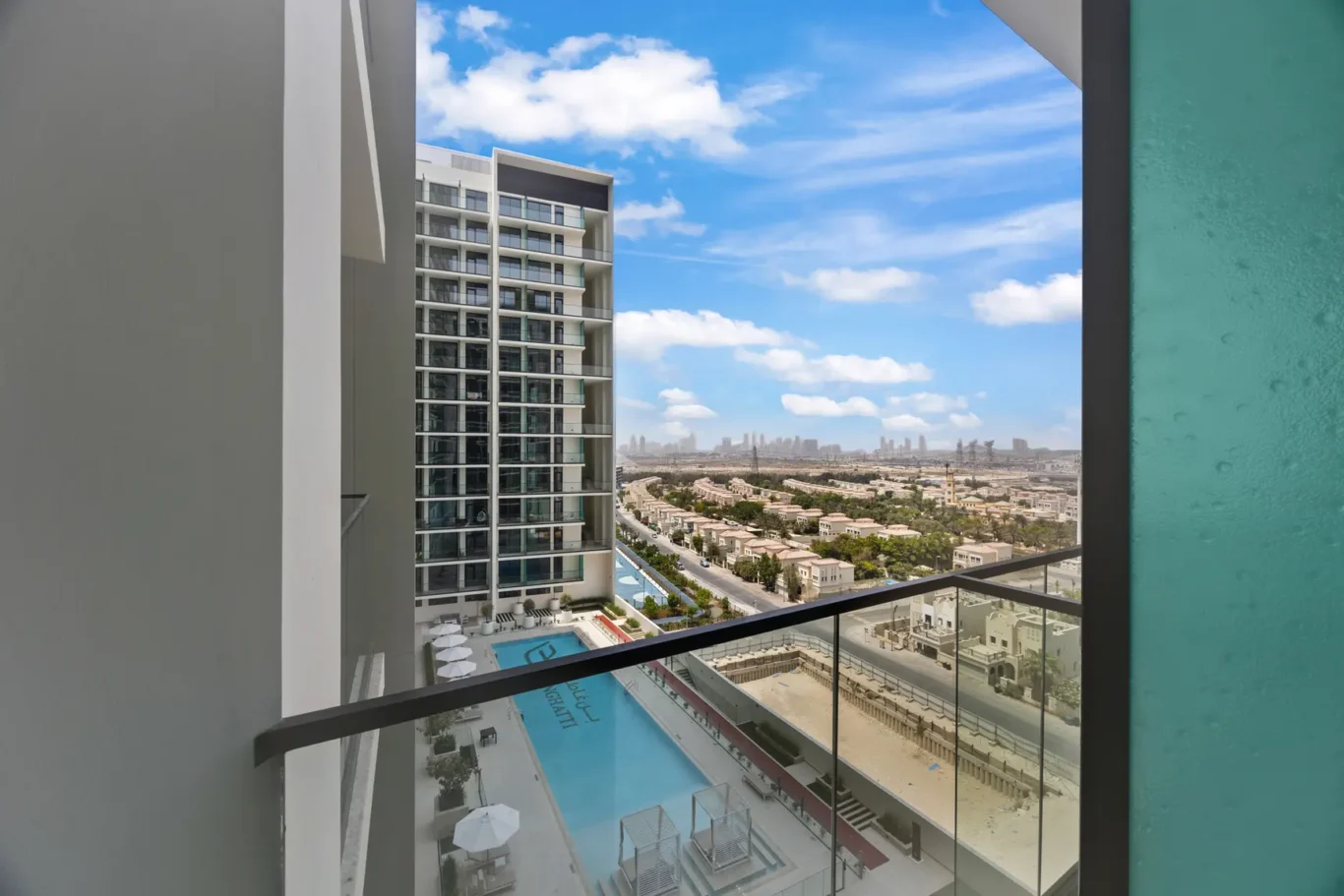 Balcony view of a modern apartment building with glass balconies and a pool below, overlooking a desert city skyline under a blue sky.