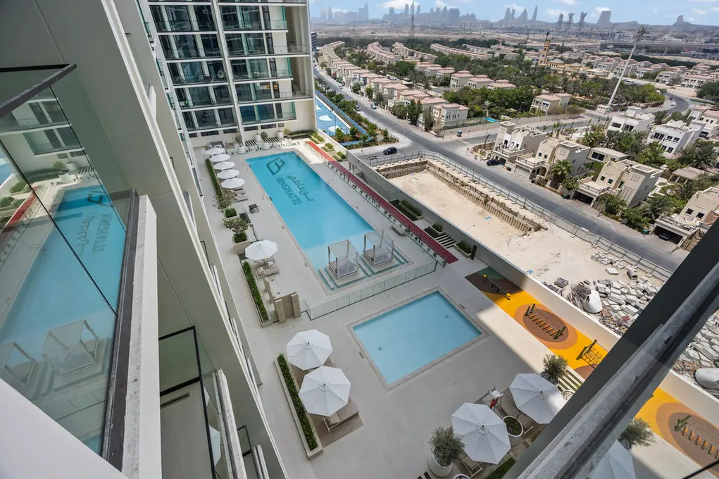 Aerial view of a modern apartment complex pool area with two blue pools, white umbrellas, and lounge chairs overlooking a suburban city.