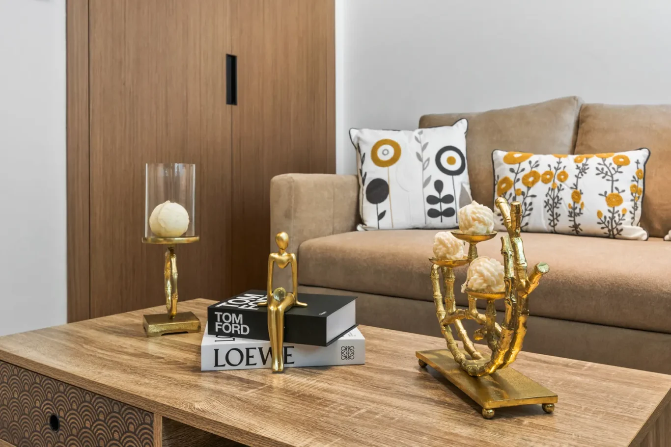Beige living room with a wooden coffee table displaying gold sculptures, stacked fashion books, and a glass cylinder candle holder beside a beige sofa with patterned cushions