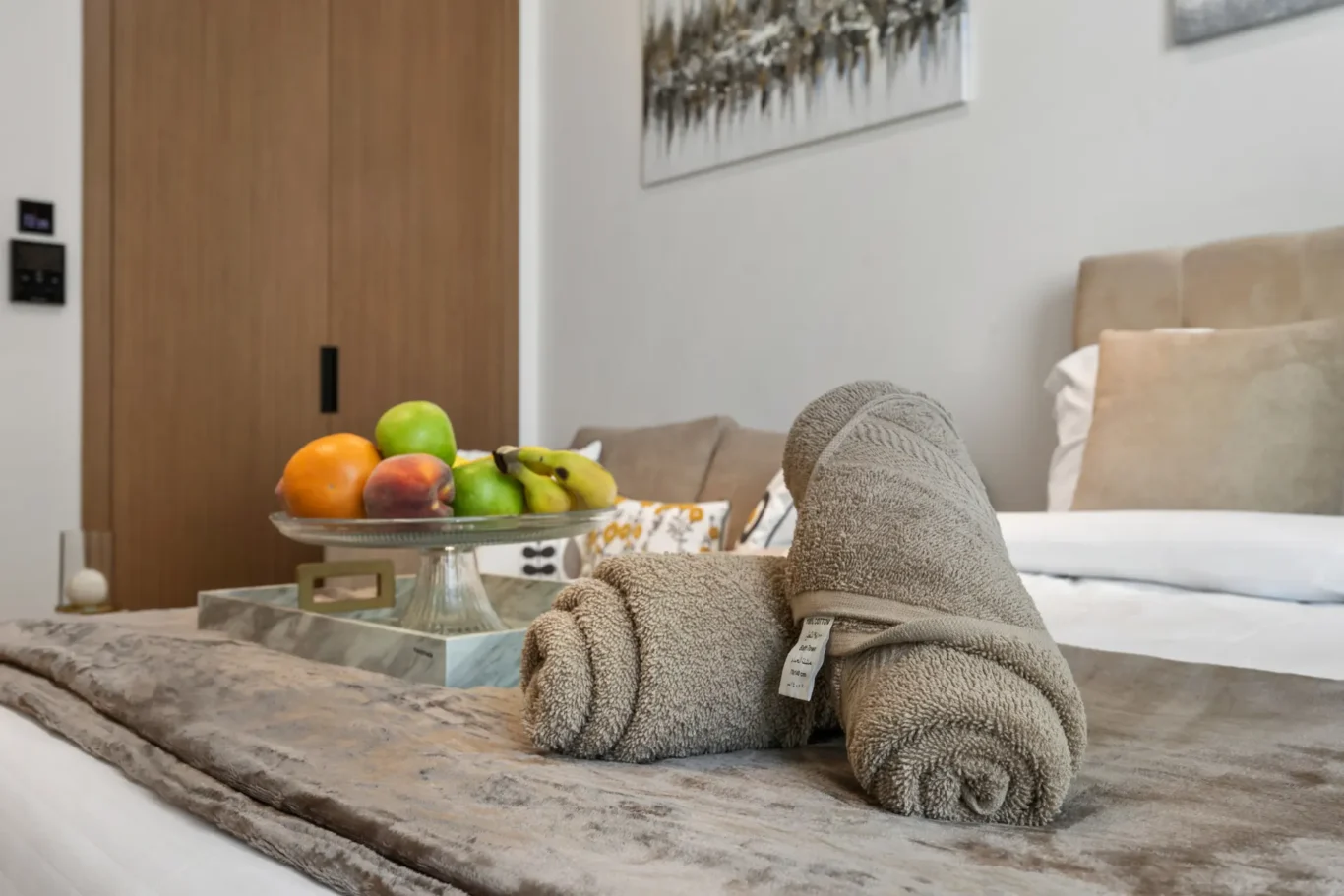 Rolled gray towels sit on a bed near a marble tray with a fruit bowl in a modern bedroom setting.
