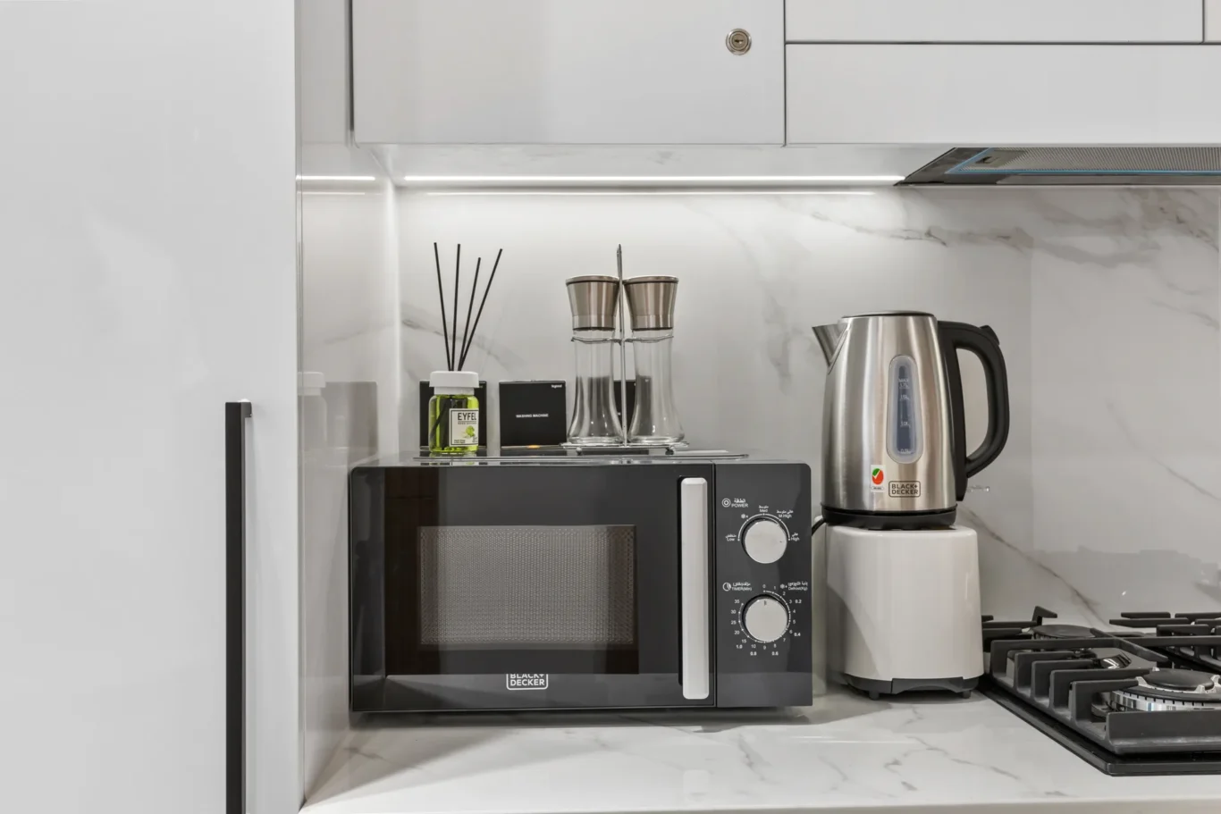 Compact kitchen counter with a Black+Decker microwave, stainless electric kettle, two glass spice jars, a reed diffuser, and a lime bottle against a white marble backsplash.