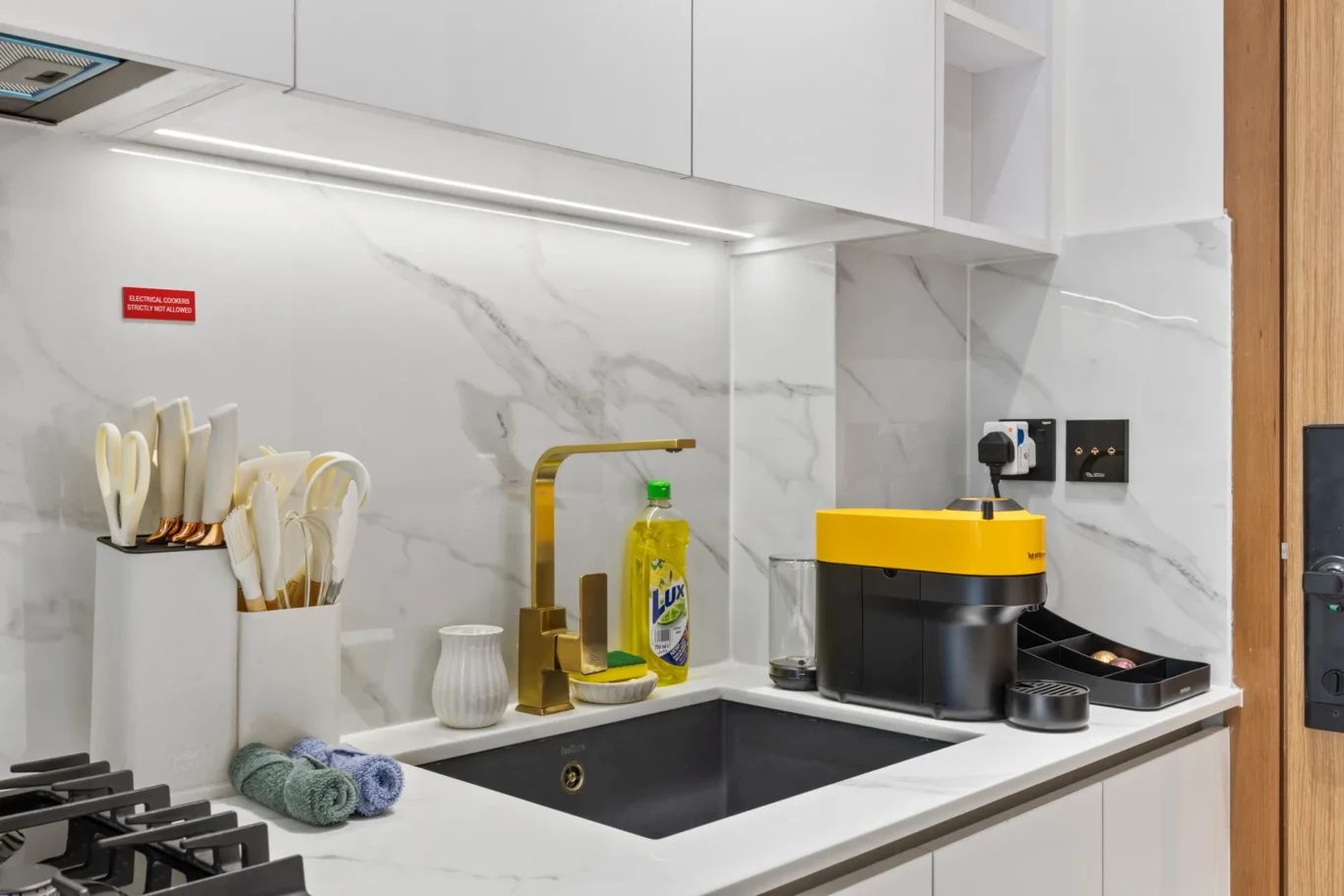 Modern kitchen sink area with a gold faucet, dish brushes, dish soap, and a yellow Nespresso machine on a marble backsplash.