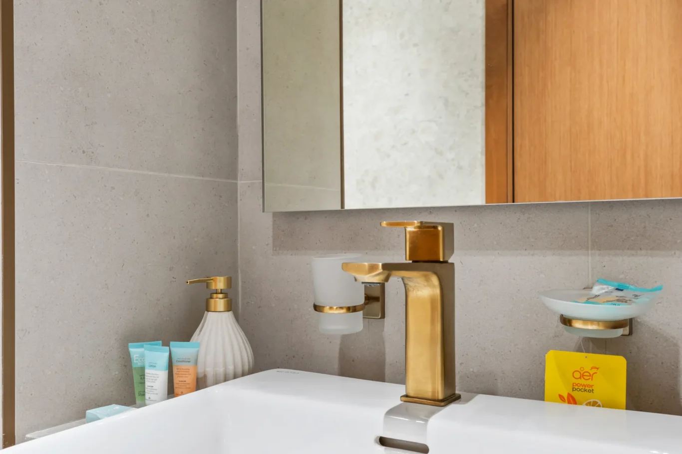 Gold faucet and white sink with beige tiled bathroom countertop and toiletries nearby.