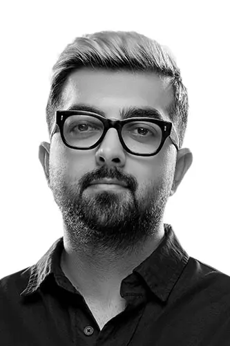 Portrait of a man with light hair and a beard, wearing thick black glasses and a dark button-up shirt, looking at the camera.