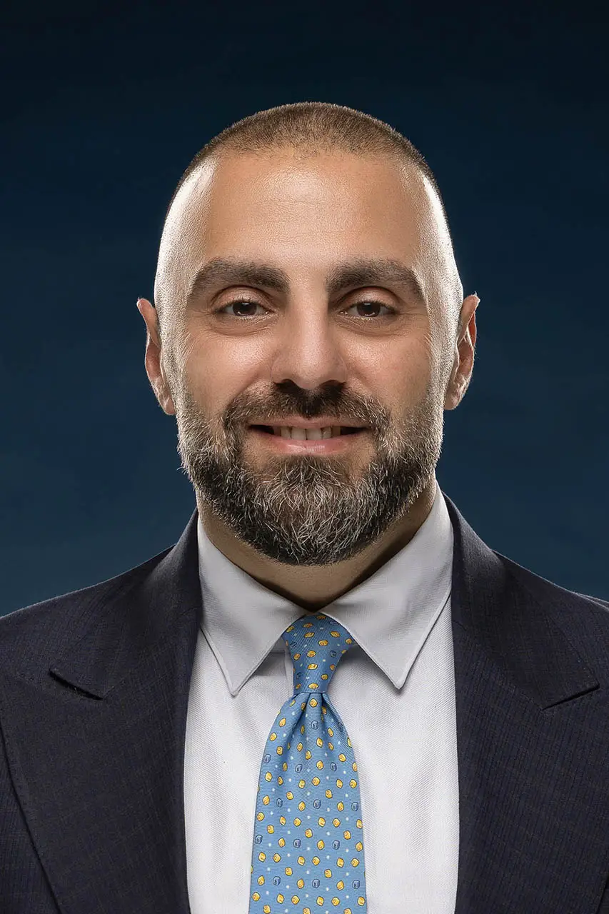 Professional headshot: man in a dark suit, light shirt, blue patterned tie, smiling against a dark gradient background.