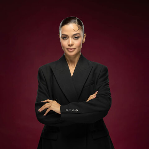 Confident businesswoman with arms crossed in a black blazer against a burgundy studio backdrop.
