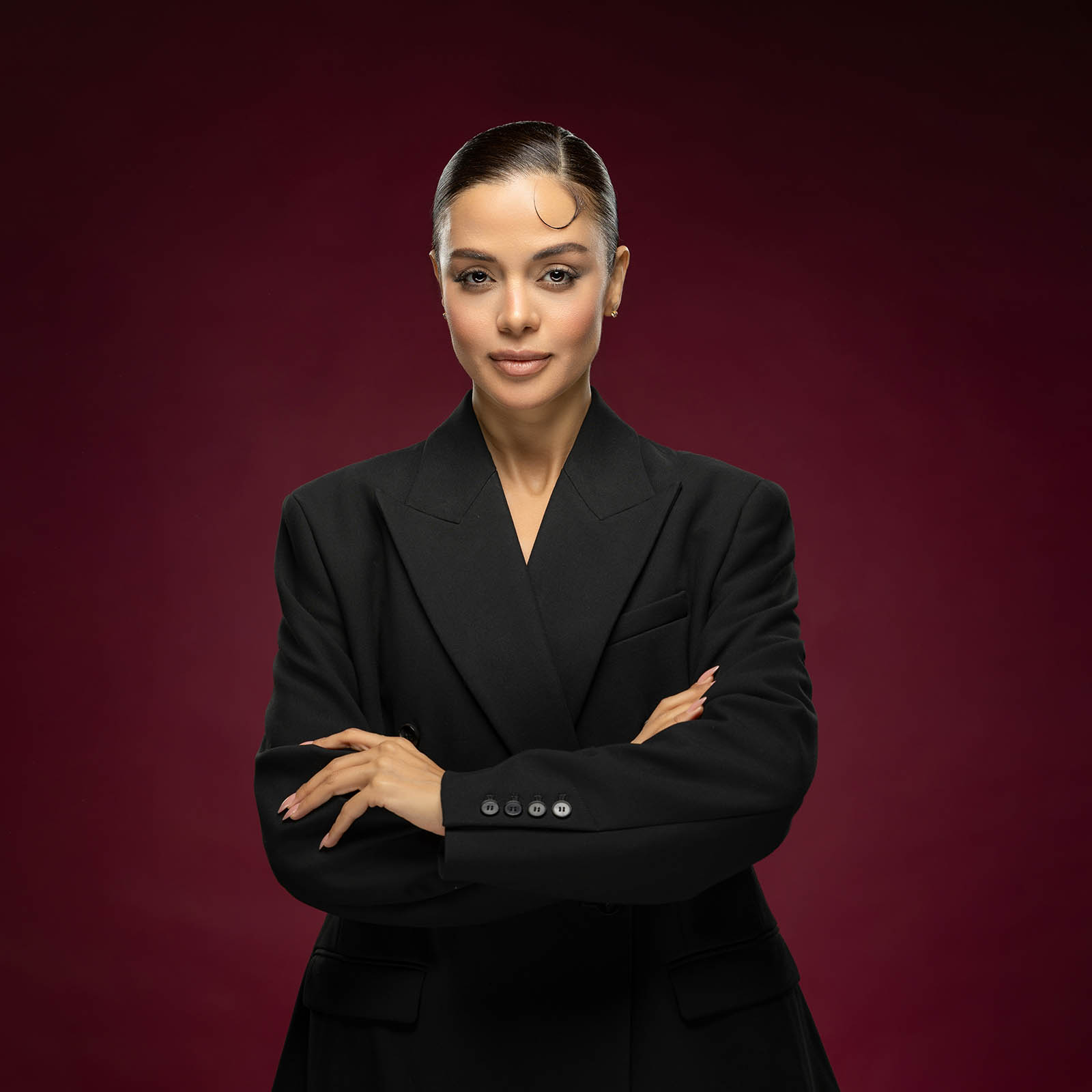 Confident businesswoman with arms crossed in a black blazer against a burgundy studio backdrop.