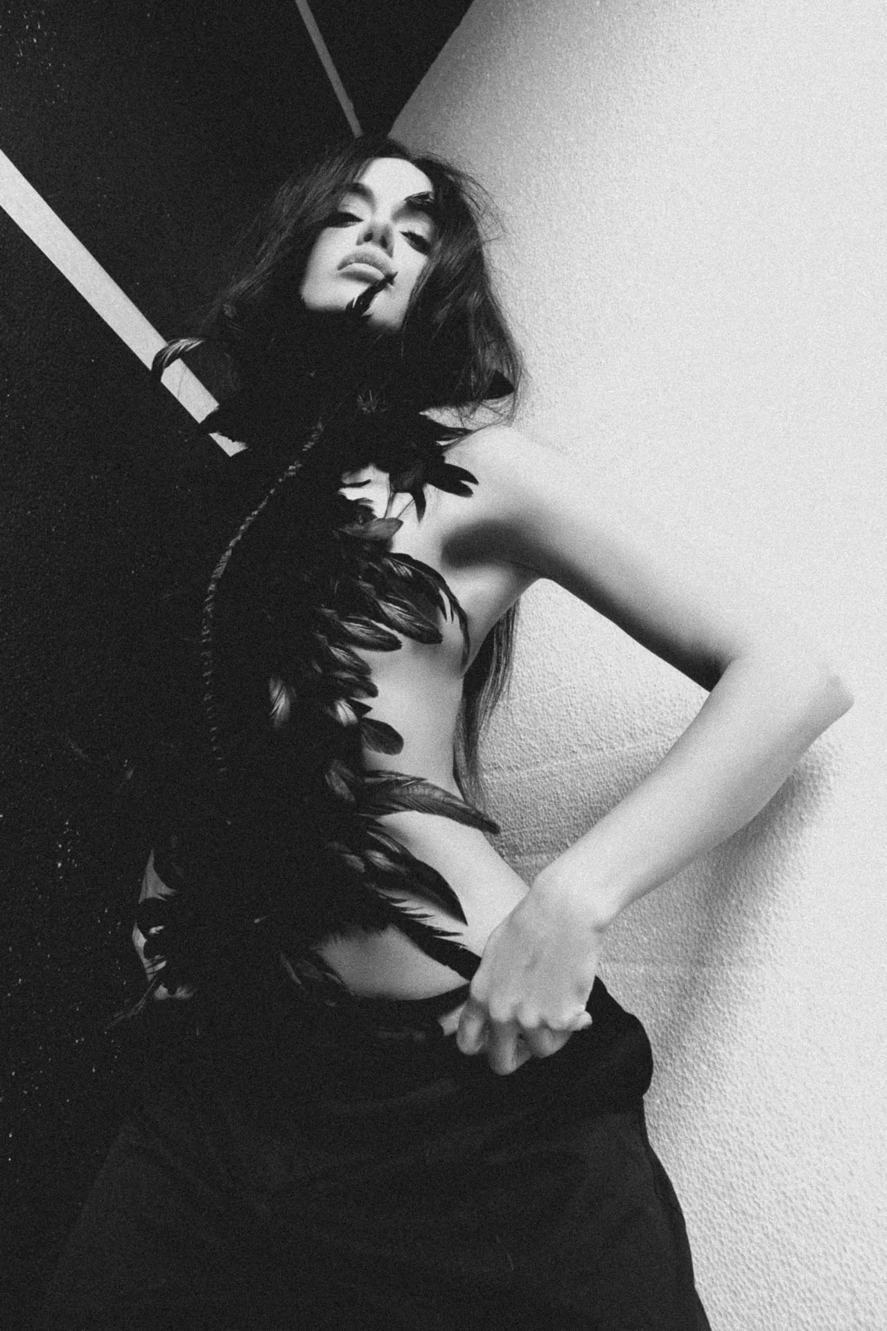 Black-and-white artistic portrait of a woman wearing a feathered adornment, leaning against a wall with one arm extended.