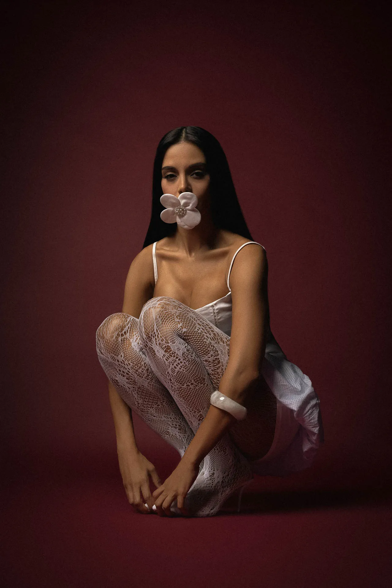 Woman seated on a burgundy backdrop with a pink flower in her mouth, wearing a white satin top and lace tights.