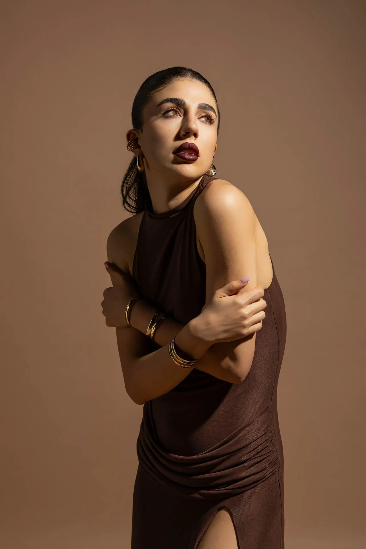 Portrait of a woman in a sleeveless brown dress, arms crossed, looking up and to the left, wearing gold earrings and bracelets against a brown backdrop.