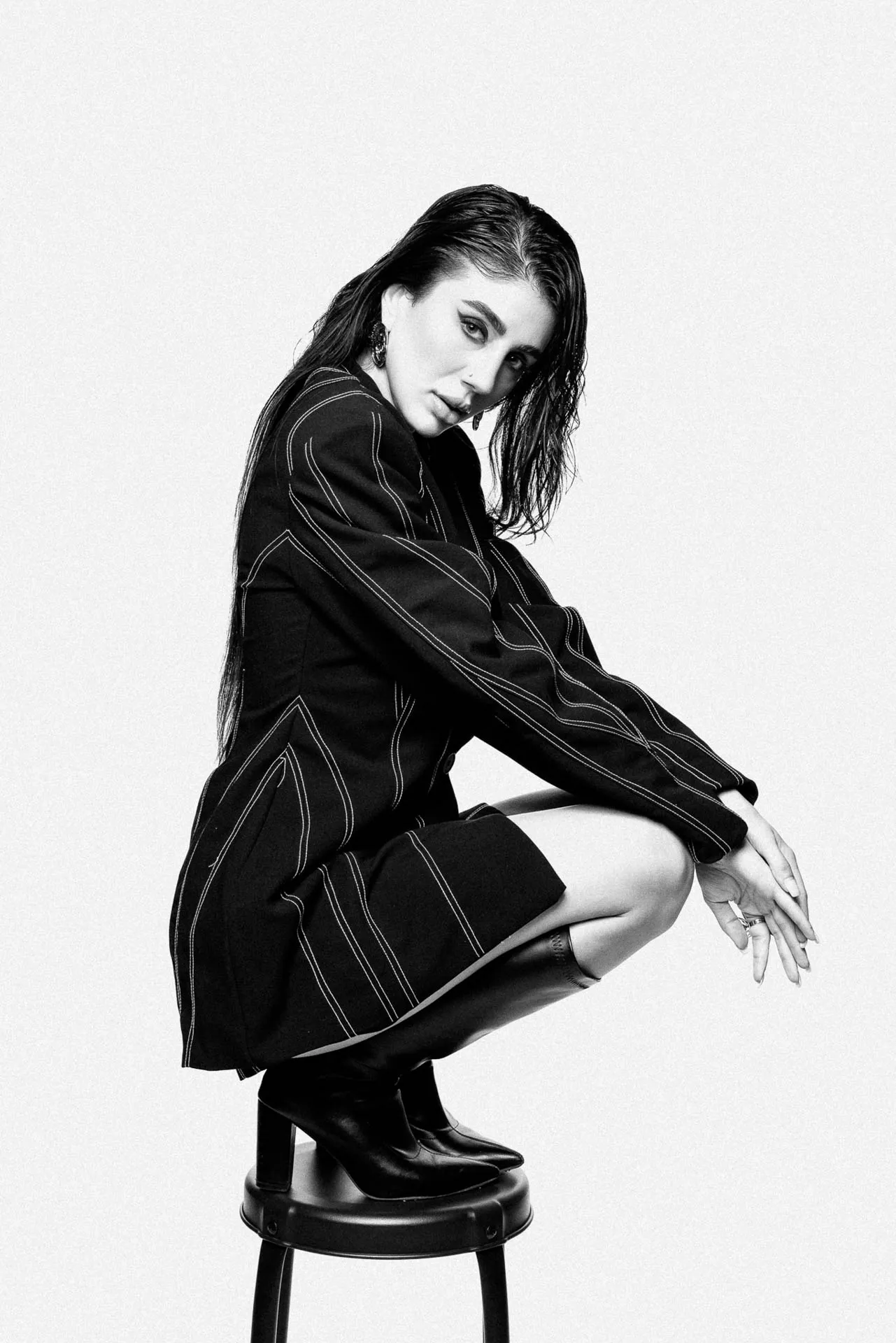 Black-and-white fashion portrait of a woman seated on a stool, wearing a pinstripe blazer and boots, looking at the camera.