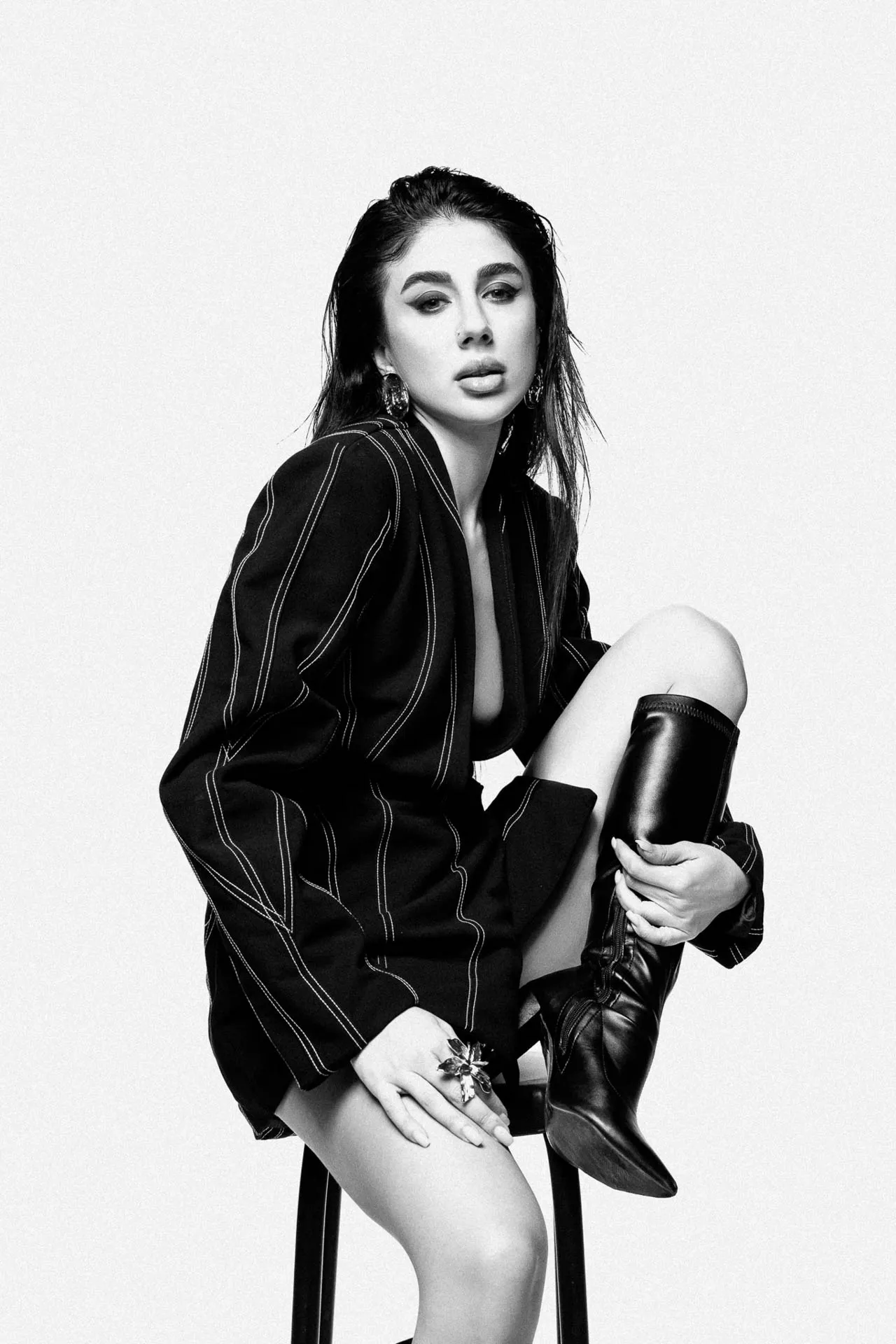 Black-and-white portrait of a woman sitting on a stool, wearing a pinstriped blazer and knee-high boot, looking at the camera.