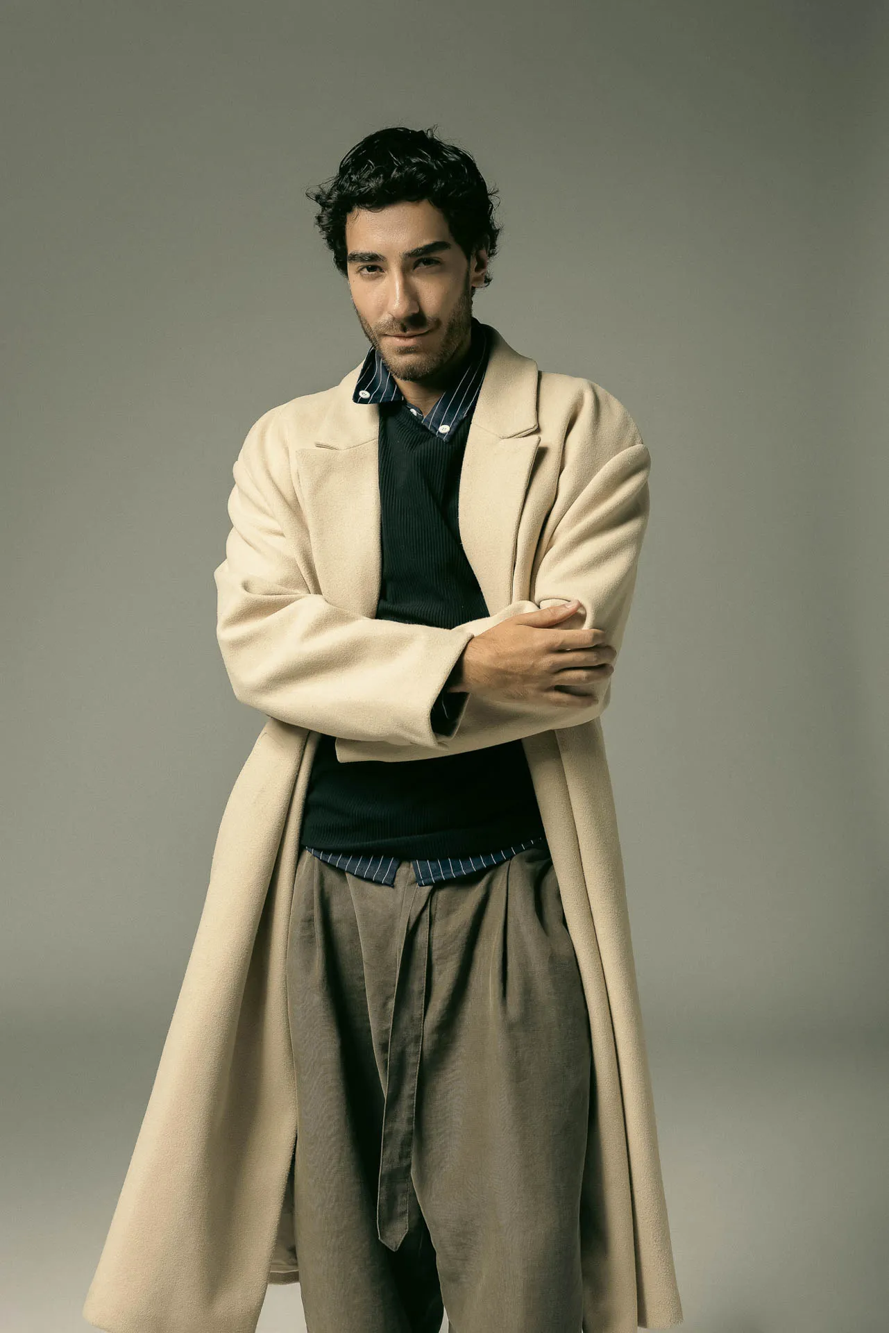 Man with dark hair wearing a cream overcoat, striped navy shirt, and tan trousers, standing with arms crossed against a gray backdrop.