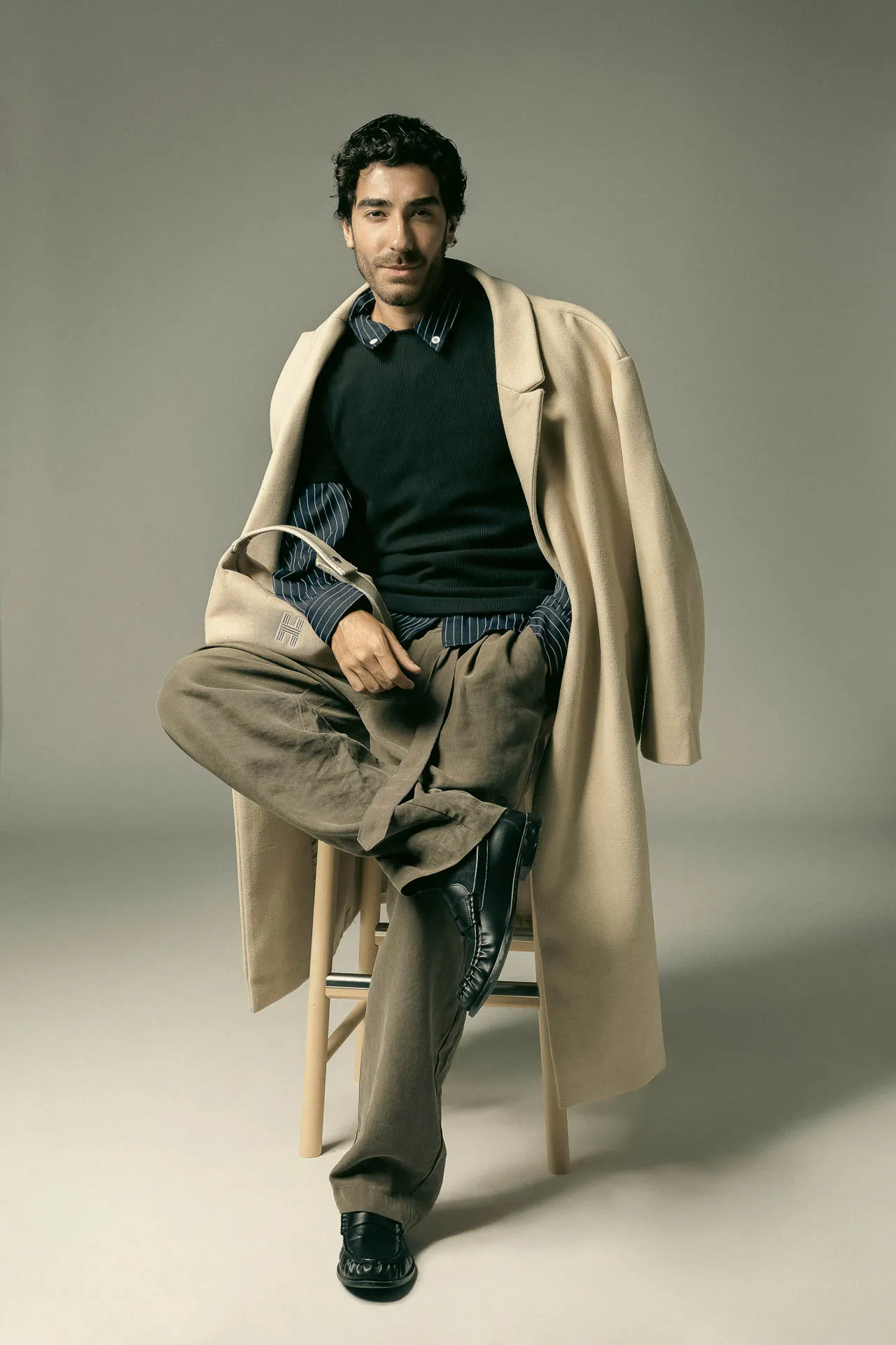 Man seated on a stool in a fashion pose, wearing a beige overcoat draped over his shoulders with a dark sweater, striped shirt, khaki pants, and black boots.