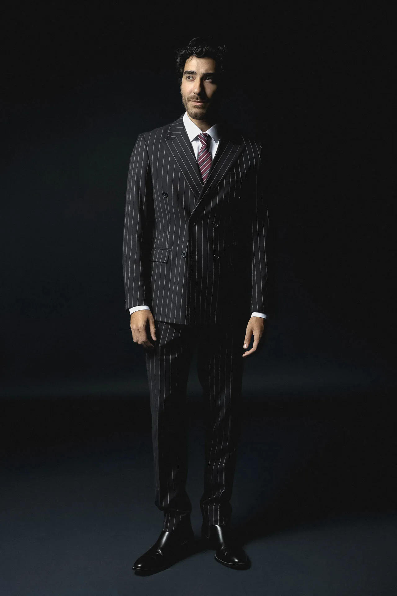 Man in a dark pinstripe suit with a red striped tie stands against a black background, looking slightly to the side.