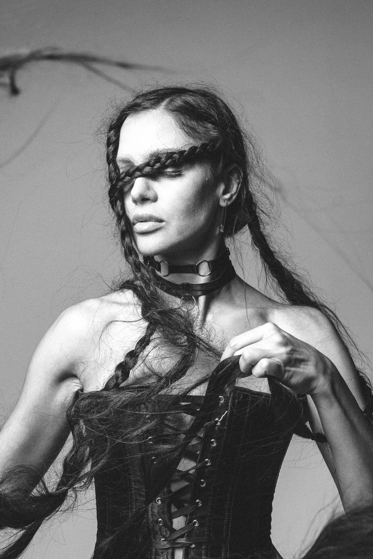 Black-and-white portrait of a person with long braided hair, wearing a leather choker and corset, looking off to the side.