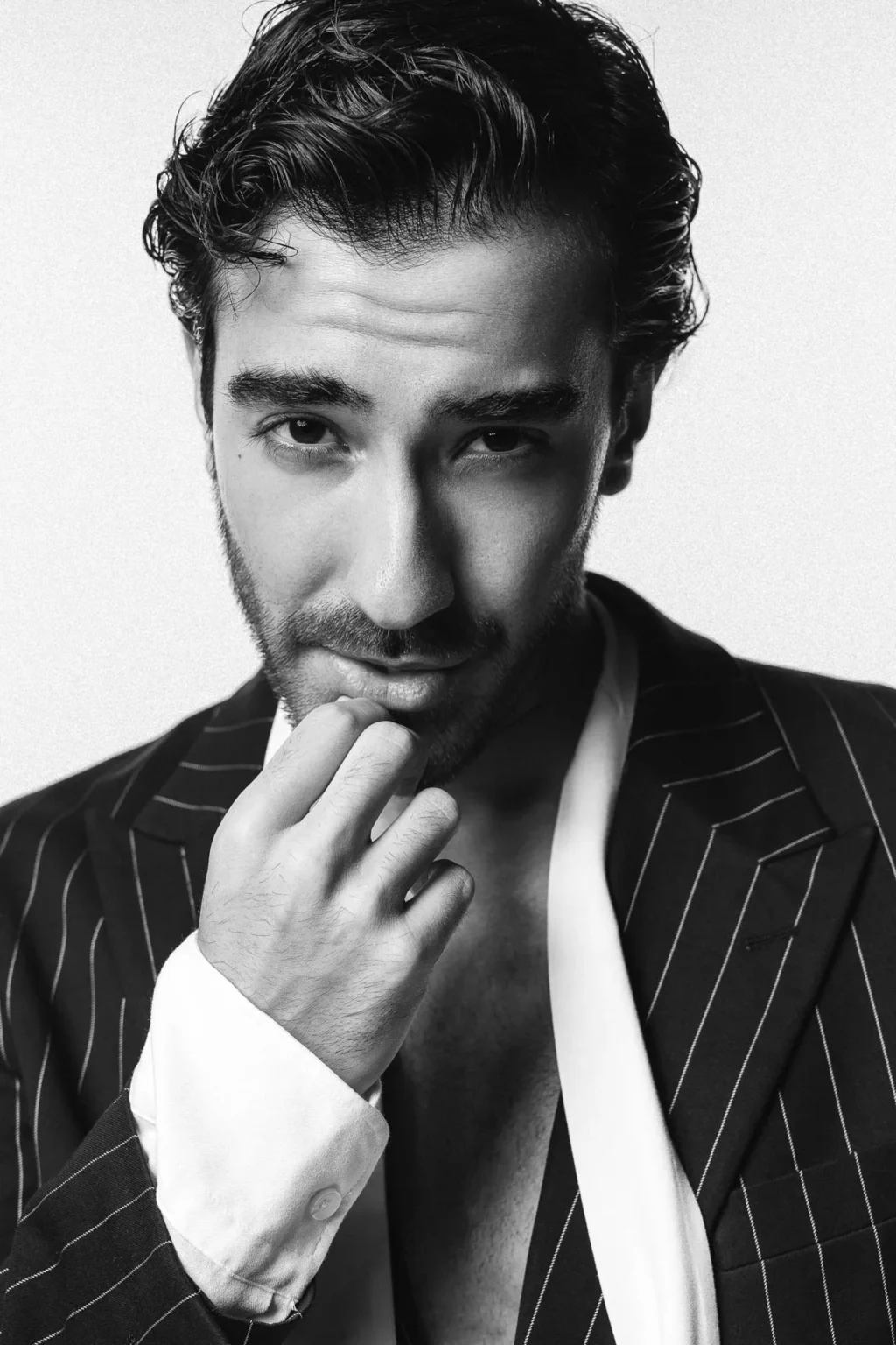 Close-up black-and-white portrait of a man in a pinstriped blazer, touching his chin with his hand and gazing at the camera.