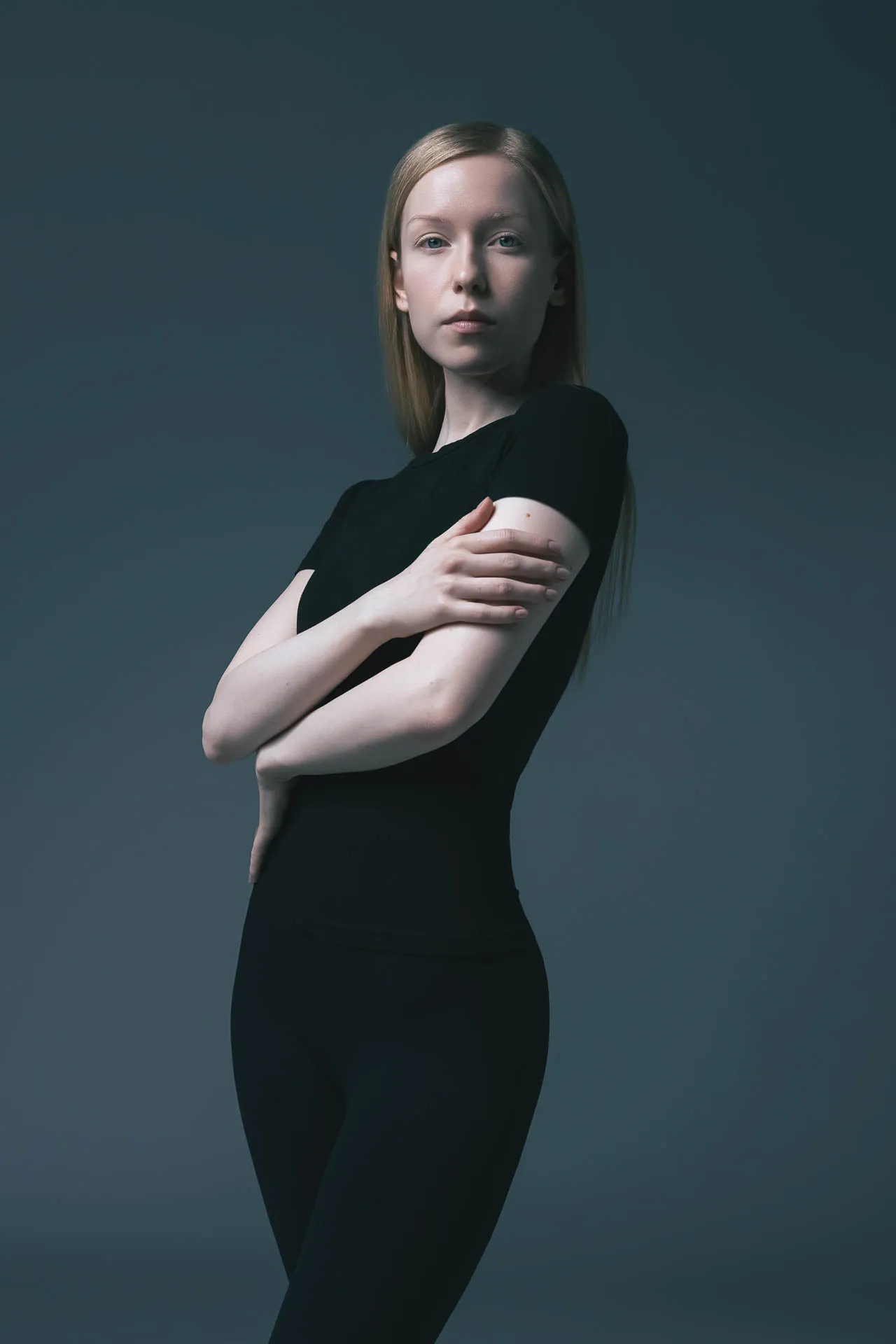 Blonde female model in a black dress, arms crossed, teal backdrop — editorial modeling portfolio Dubai studio
