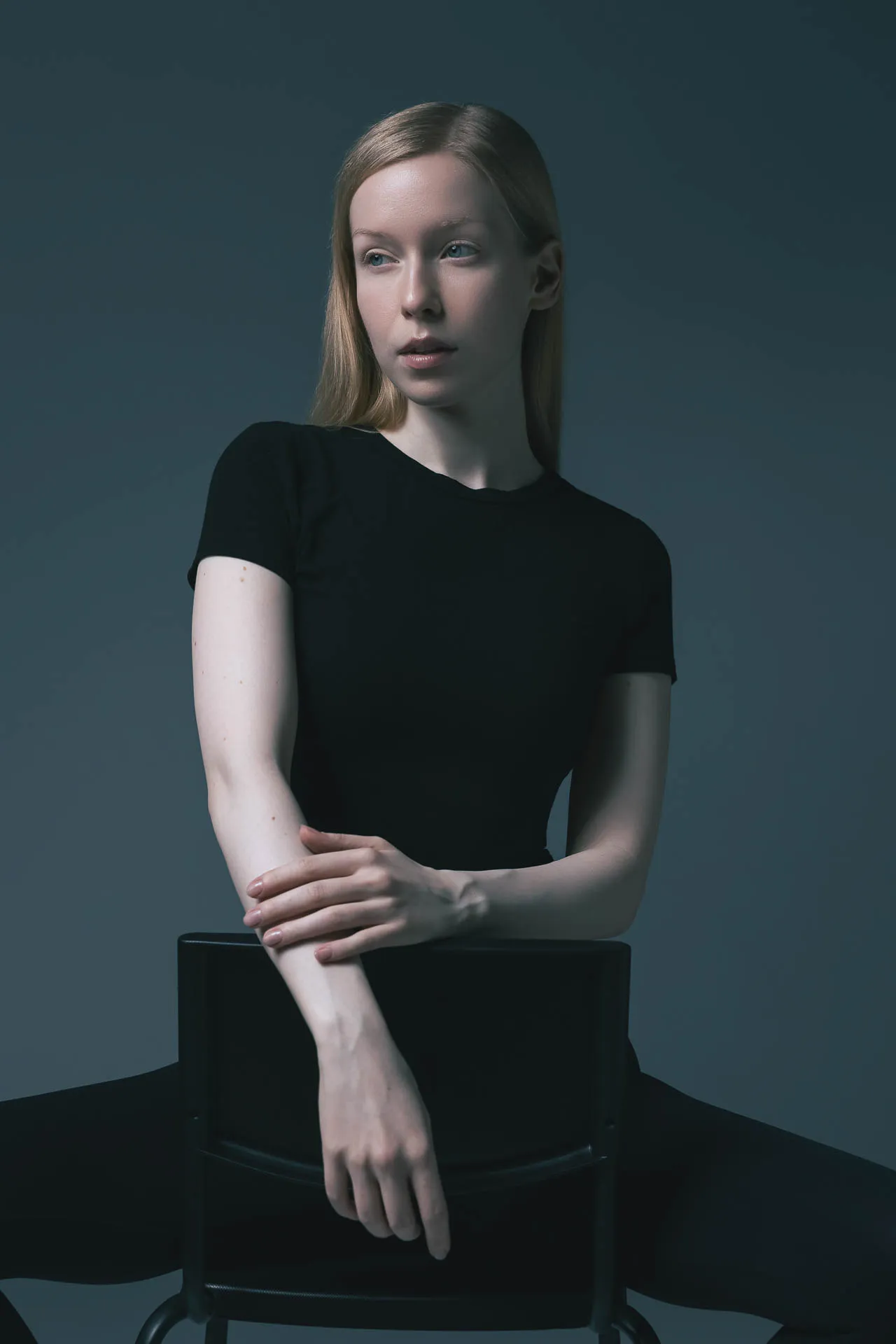 Young woman with blonde hair wearing a black top, seated with arms crossed on a chair against a dark teal background, looking to the side.