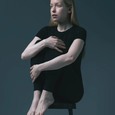 Blonde female model in black, arms crossed, seated on a stool against a teal backdrop — Dubai modeling portfolio
