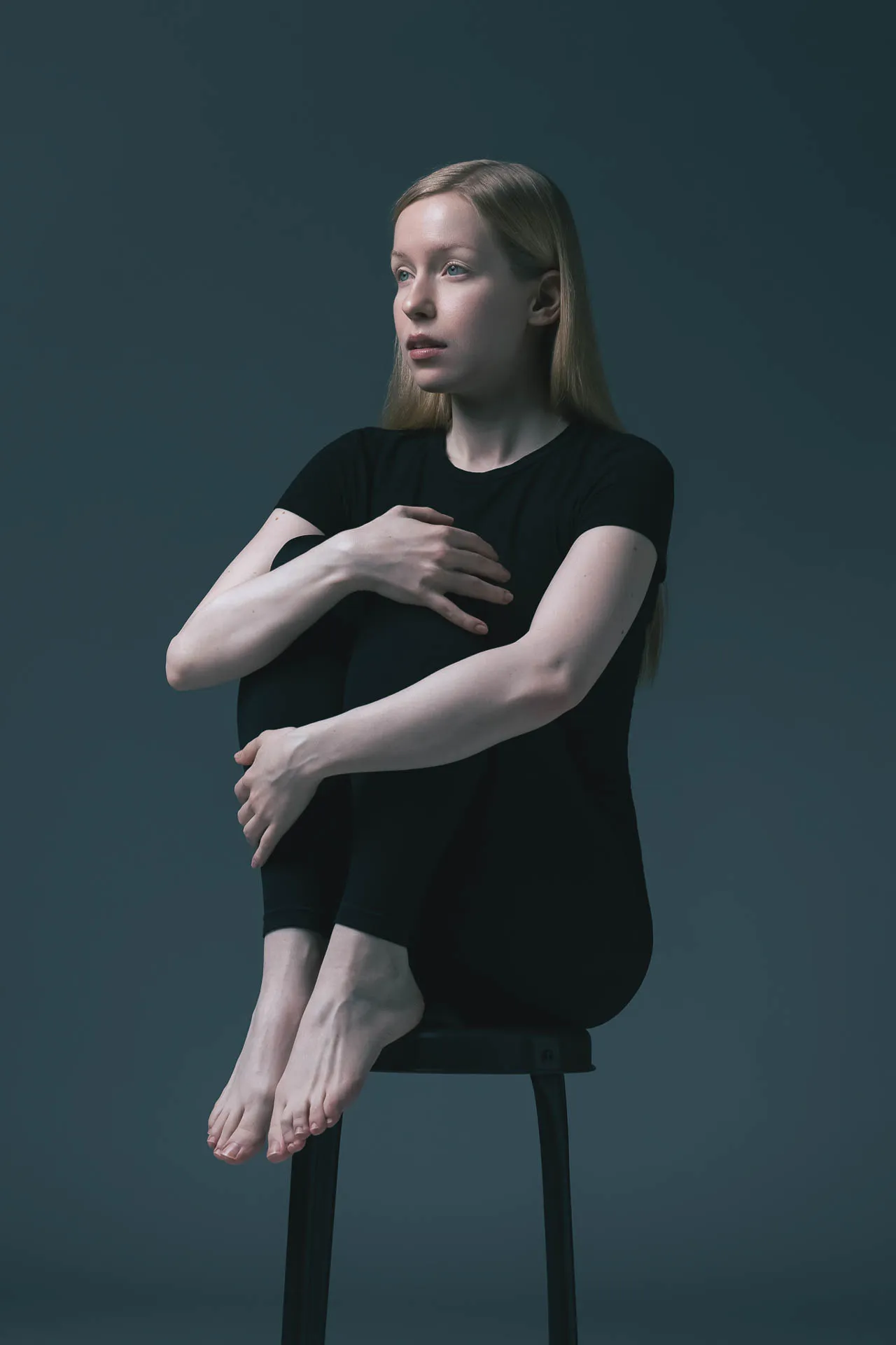 Blonde female model in black, arms crossed, seated on a stool against a teal backdrop — Dubai modeling portfolio