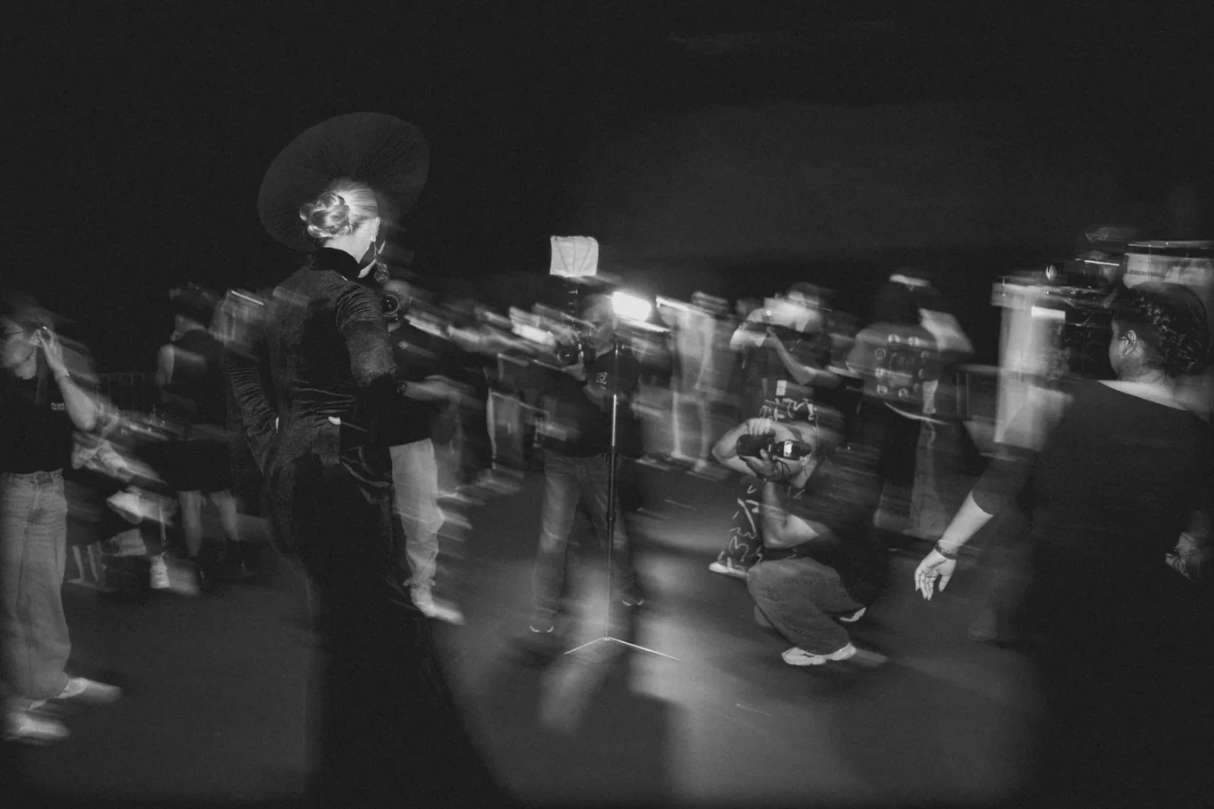 Woman wearing a large round hat stands with her back to the camera as a crowd moves past at night, captured with long exposure creating motion blur.