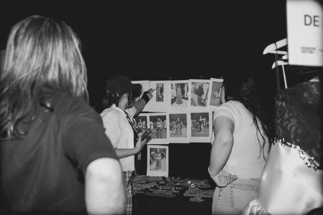 People gathered at an event looking at a wall of fashion photos pinned to a display board at night.