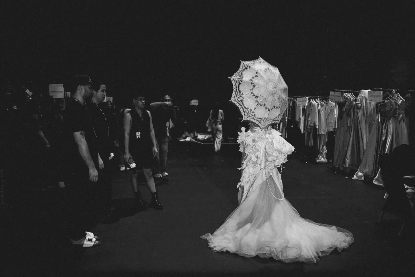Backstage at a fashion show: a model in an elaborate white gown with a lace parasol walks past racks of gowns while crew watches.