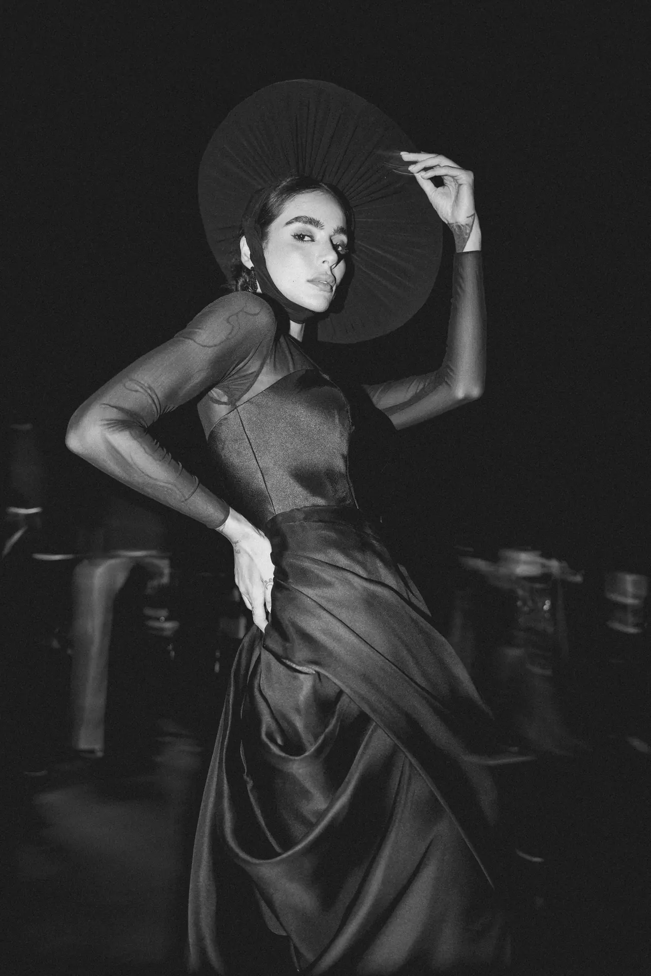 Black-and-white photo of a female model in a satin gown with a large round hat, posing with hand on hip.