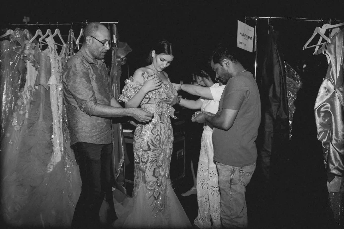 Backstage dressing room: a woman in a sequined, ornate gown is being adjusted by two men while other staff help with outfits nearby beside clothing racks.