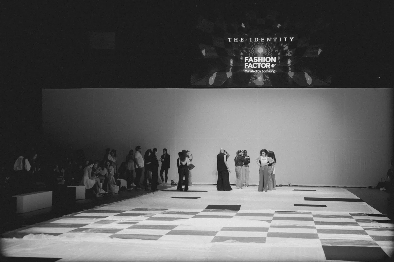 A fashion show on a lit runway with models posing while a seated audience watches from the sides.